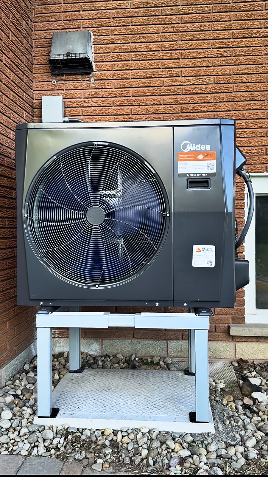Midea heat pump outdoor unit on a raised steel stand against a brick wall, with a Kelvin Mechanical service sticker