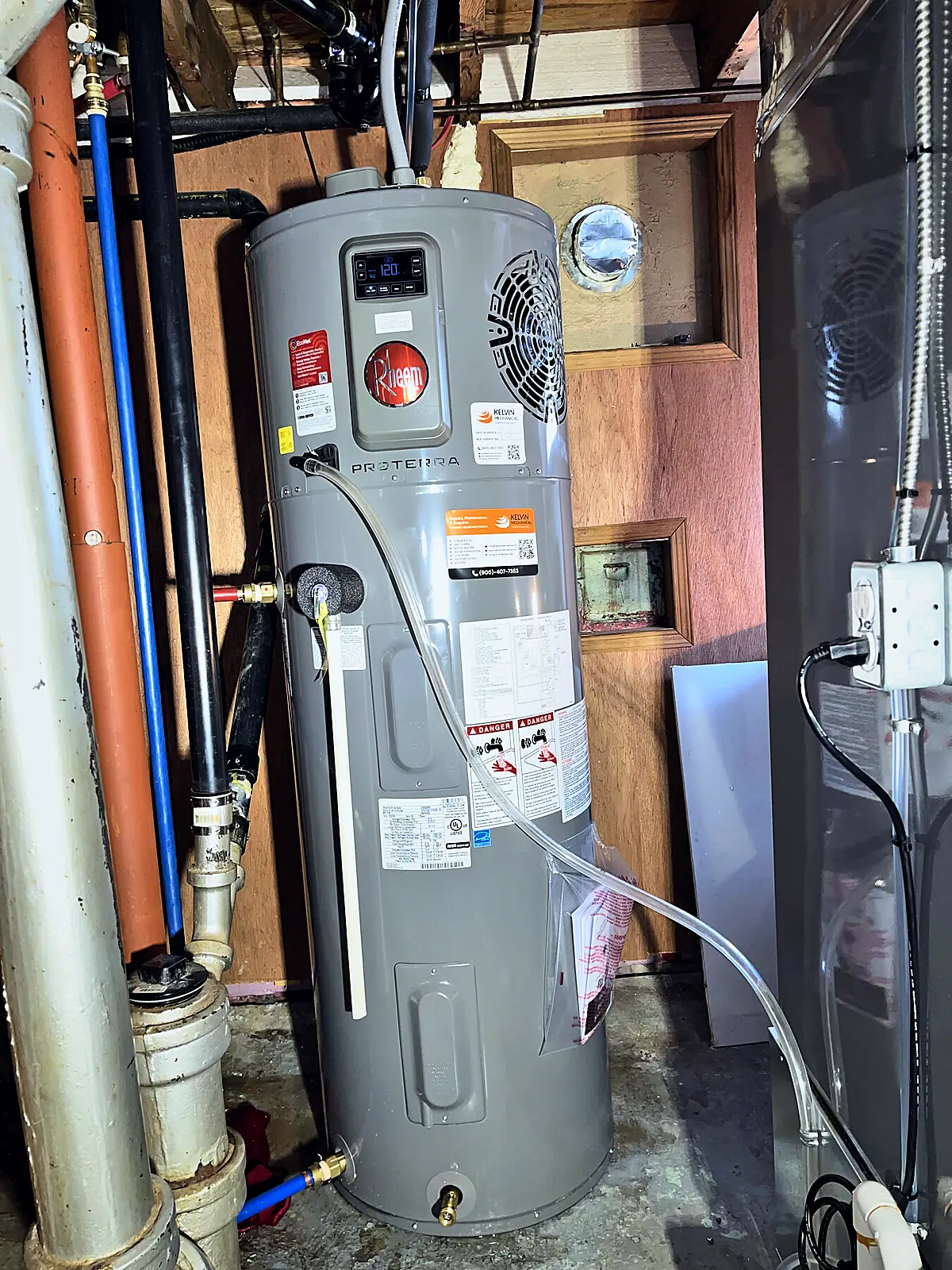 Rheem heat pump water heater with its water piping and clear condensate line in a basement utility room
